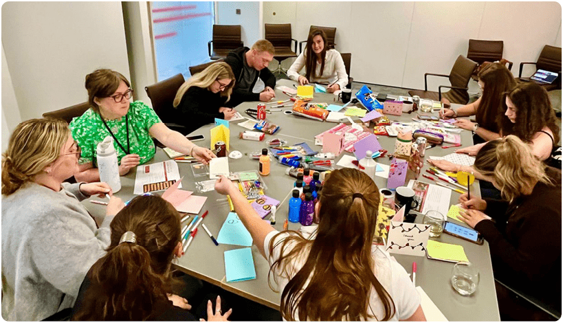 A group of people with various hairstyles are seated around a large table covered with art supplies, papers, and snacks, actively engaged in a creative or collaborative activity in a bright room. Description generated by AI