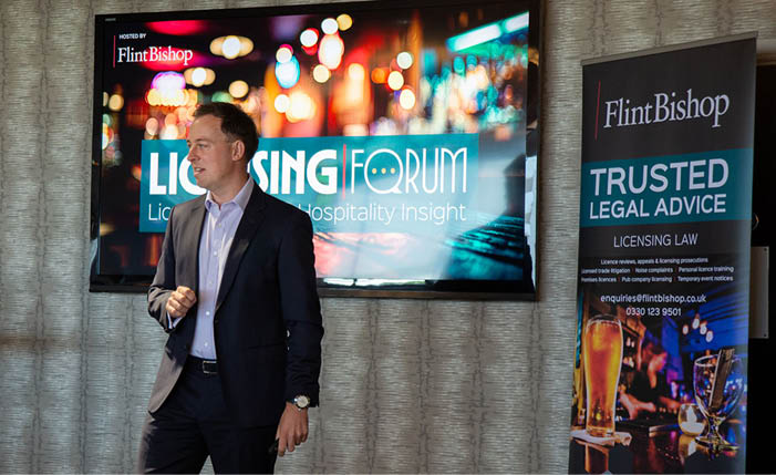 A person in a suit is standing and speaking in front of a large screen and a banner displaying legal advice information in a professional indoor setting. Description generated by AI